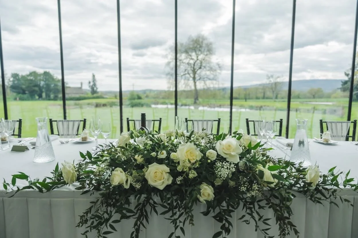 Elegant wedding table setup with white roses and green foliage centerpiece, glassware, and a scenic outdoor view through large windows.