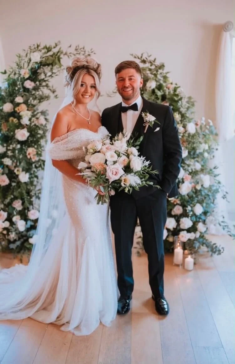 A bride and groom on their wedding day, standing in front of a broken floral arch, candles on the floor, both smiling and holding a bouquet of flowers. The bride is wearing a white wedding dress and the groom is in a black tuxedo.