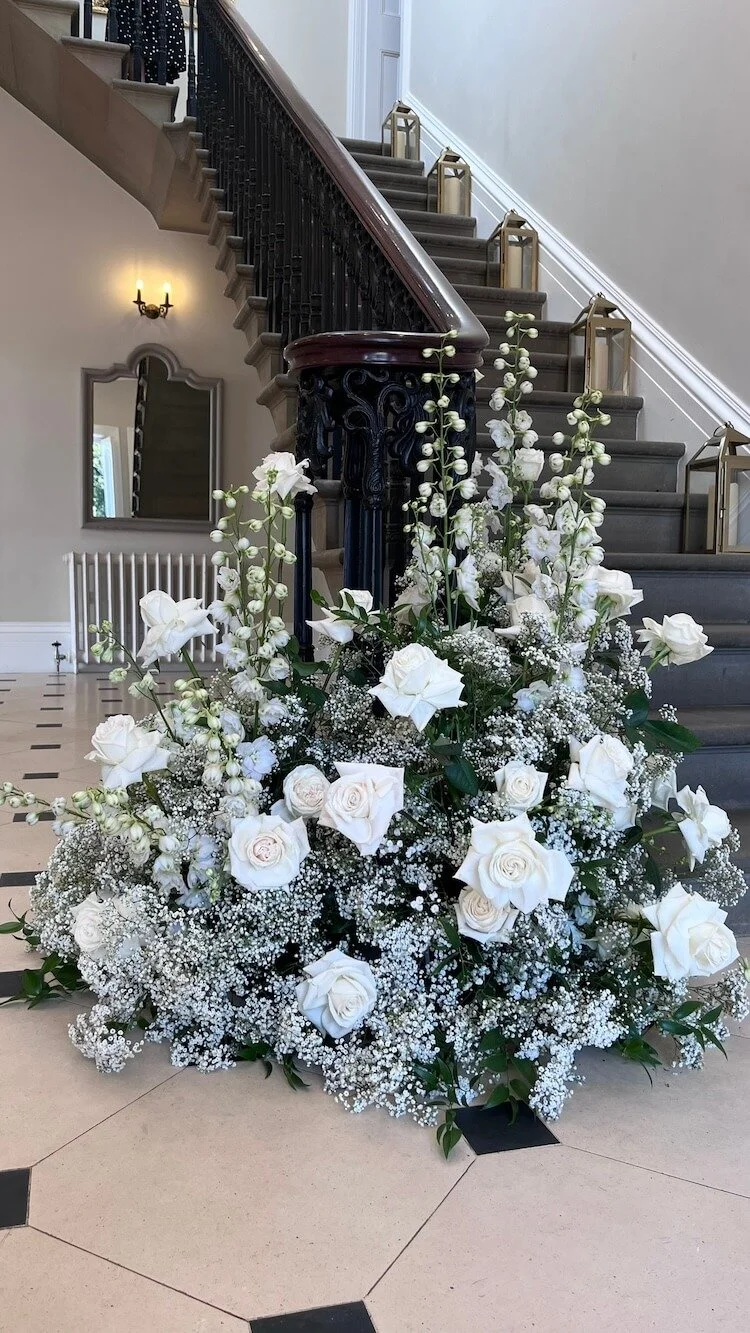 A large arrangement of white roses, baby’s breath, and tall white flowers in front of a staircase inside a building with beige walls and black and white tiled floor.