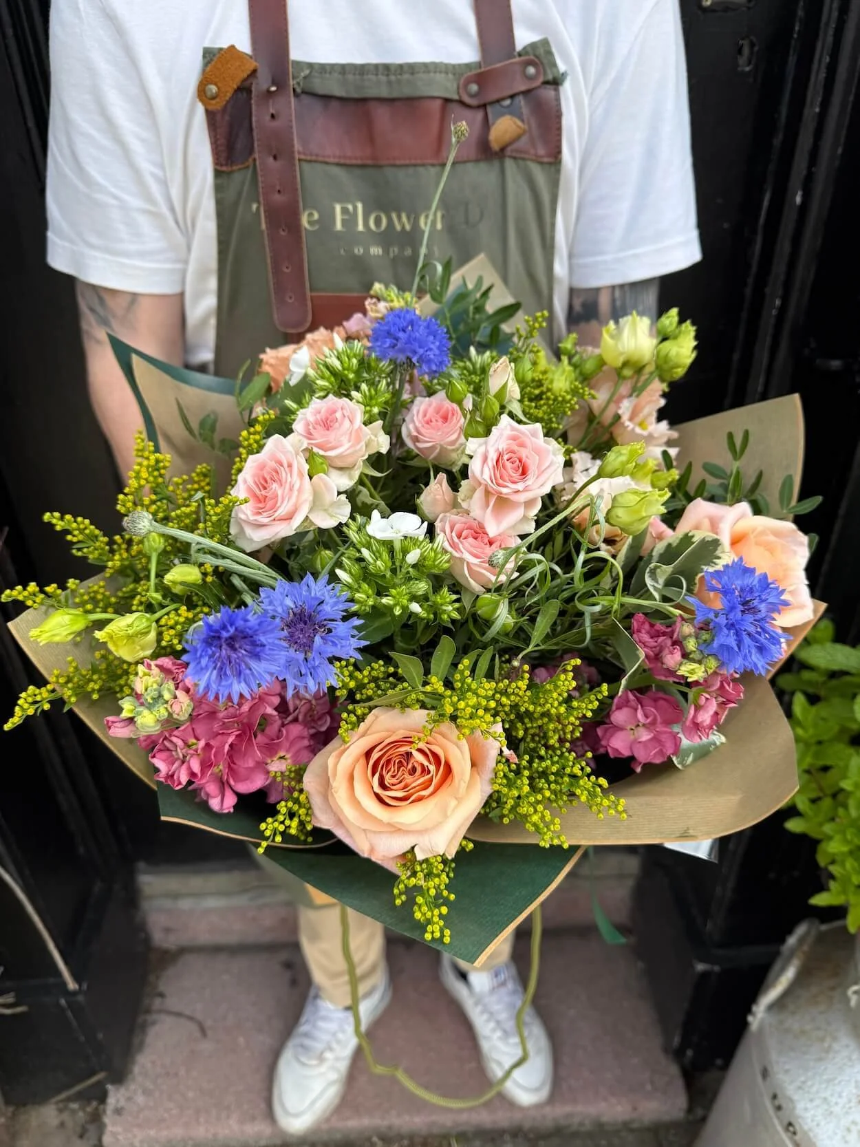 Person wearing an apron holding a vibrant bouquet of flowers, including light pink roses, blue cornflowers, and yellow accents, standing on a doorstep.