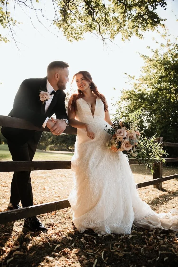 A bride and groom leaning on a wooden fence outdoors, with trees and sunlight in the background. The bride is holding a bouquet of flowers and has long red hair, while the groom is dressed in a black tuxedo with a boutonniere. They are smiling and lo
