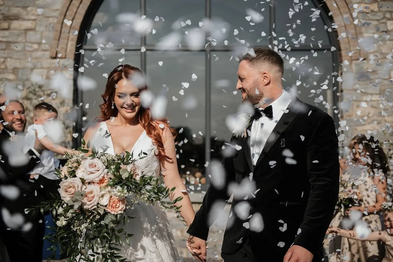 Bride and groom smiling and holding hands as confetti falls during their wedding ceremony outside in front of a stone building.