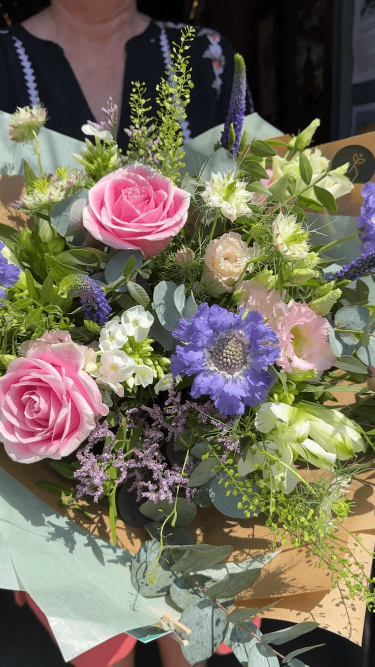 A colourful gift-wrapped bouquet of pink roses, purple flowers, white flowers, and green foliage held by a person in a black top.