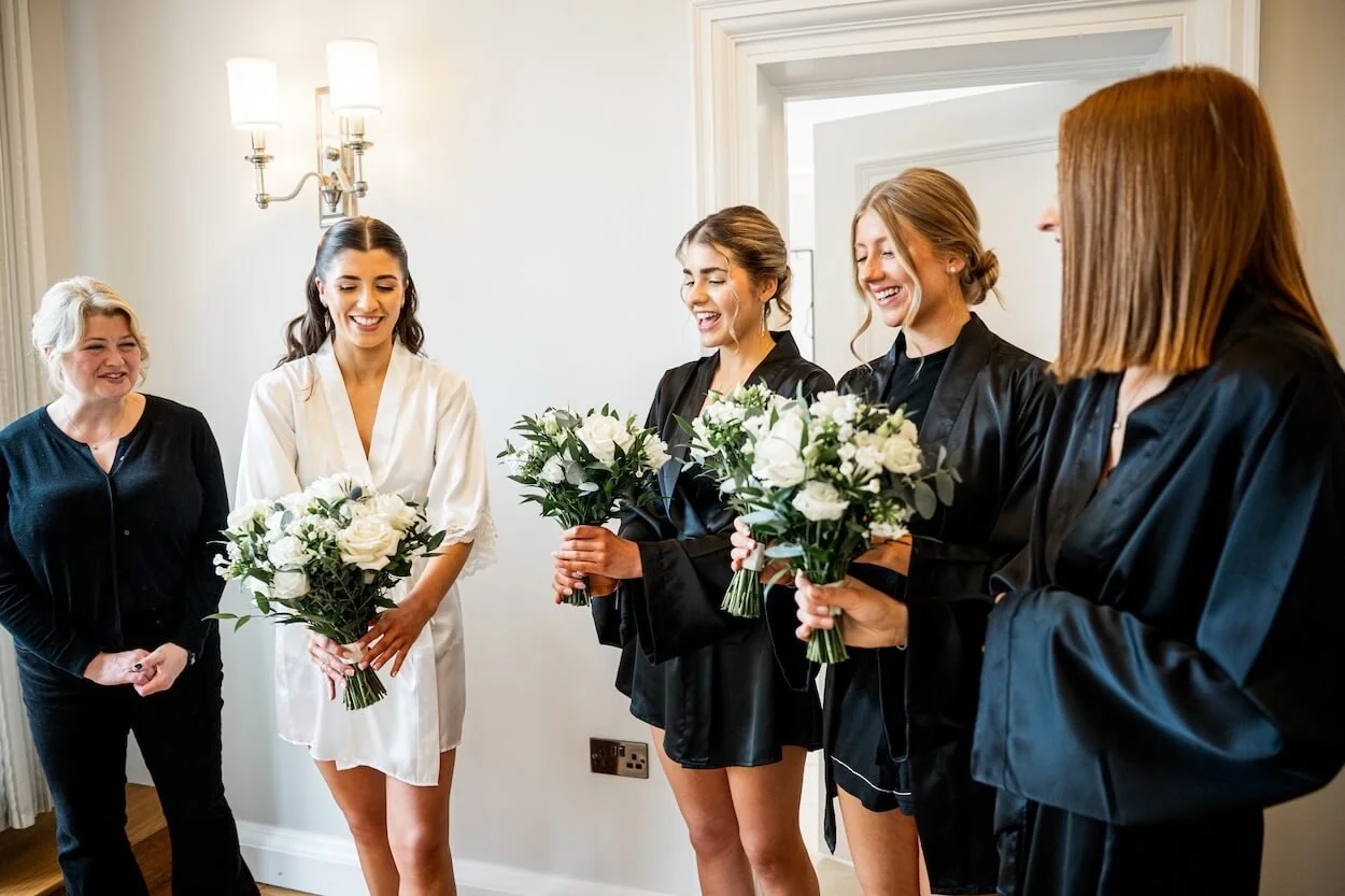 Group of five women dress in satin robes, four holding bouquets of white and green flowers, gathered inside a room, looking at their bridal party bouquets.