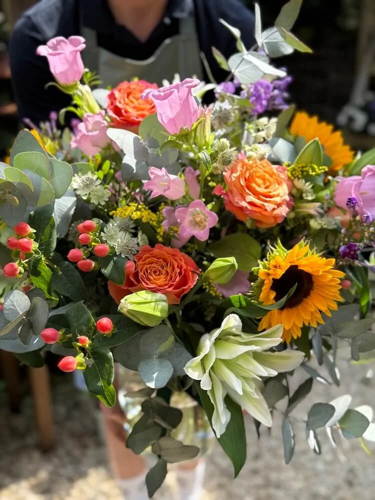 A person holding a bright and colourful bouquet of flowers including sunflowers, roses, pink bell-shaped flowers, white lilies, and greenery.