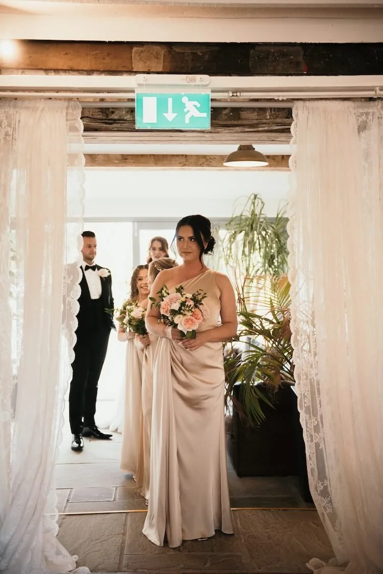 Bridal party walking through lace curtain doorway, holding bouquets, during a wedding preparation or ceremony.