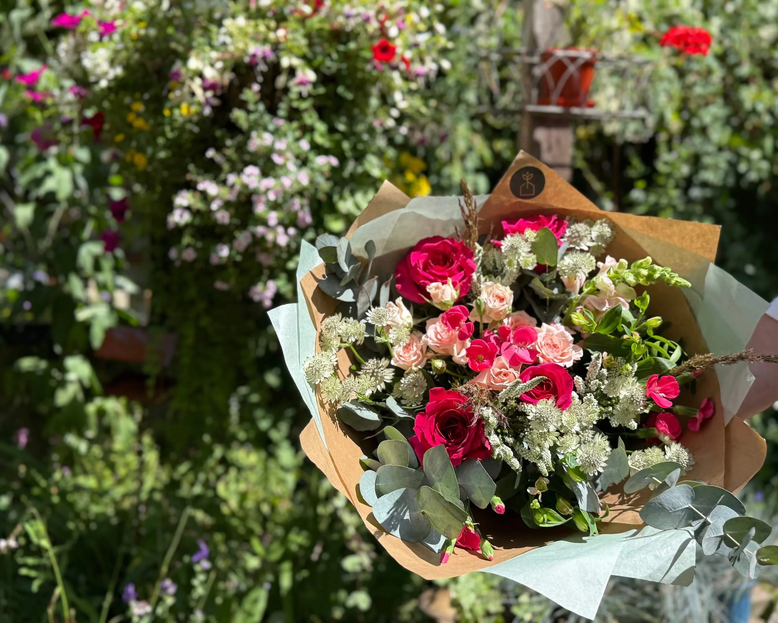 A mixed bouquet, including hot pink roses, soft pink flowers and white filler flowers, wrapped in brown paper with sage green tissue paper, held outdoors in front of a garden with flowering plants.