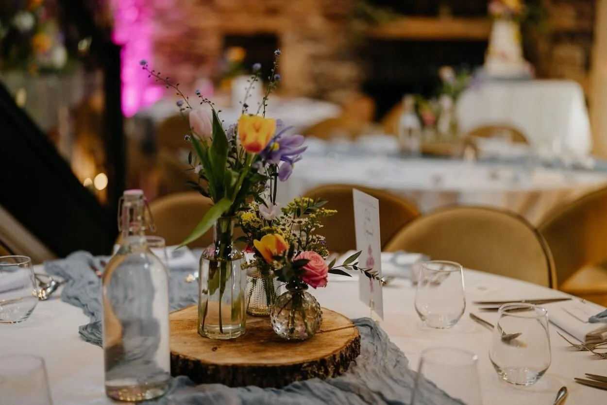 Table decorated with a centerpiece of flowers in glass bud vases on a wooden slice, surrounded by water bottles, glasses, and cutlery at a wedding reception with warm lighting and blurred background.