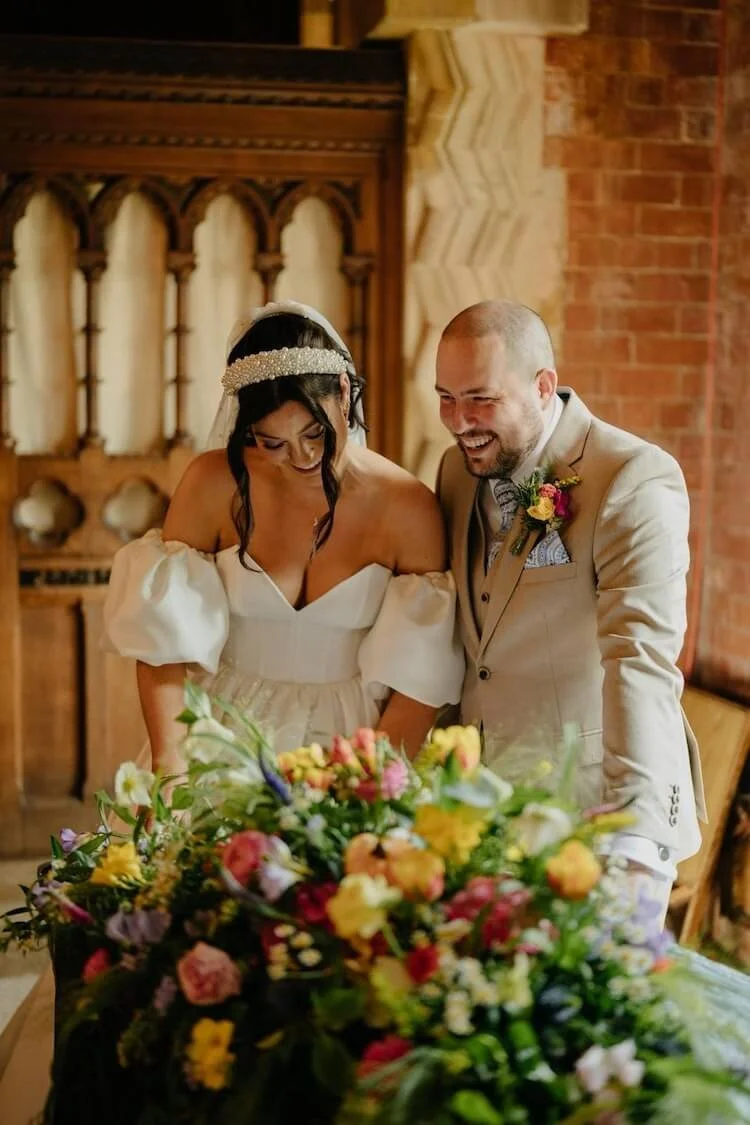 A bride and groom at their wedding, smiling and looking at a colourful registrar table arrangement of flowers.