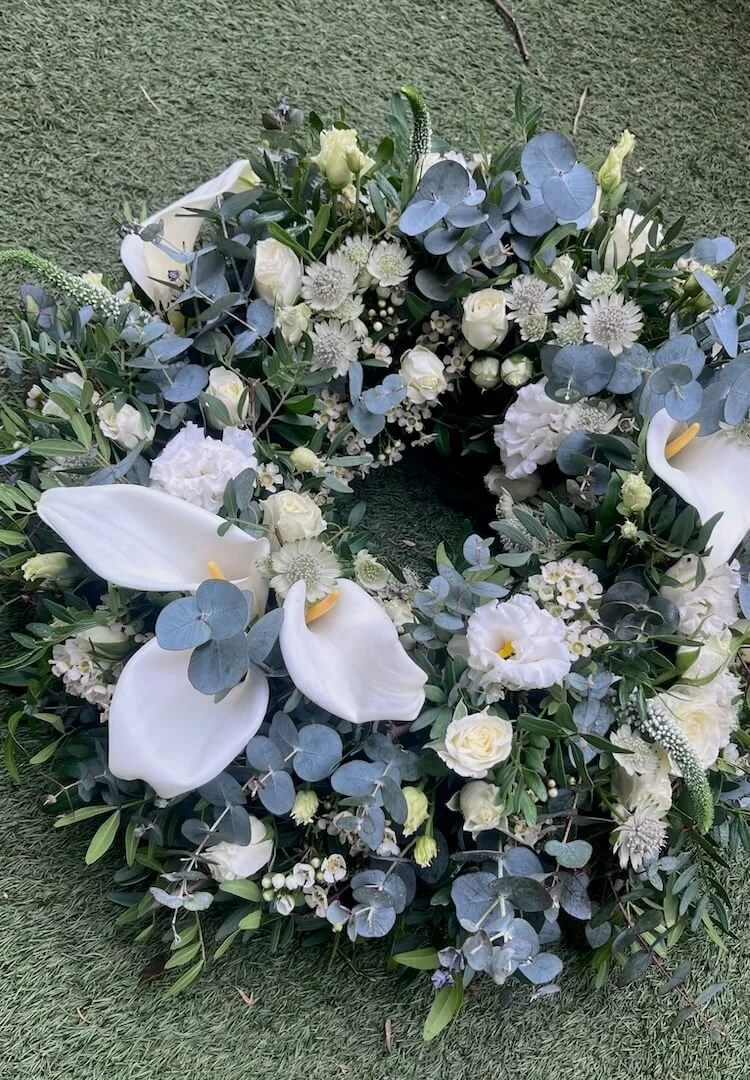 Wild, garden-style sympathy floral wreath with a mix of flowers and foliage, including white calla lilies and eucalyptus, on an artificial grass surface