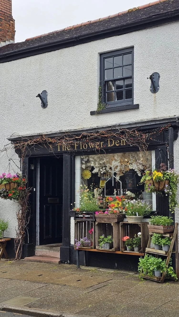 Front of a flower shop named 'The Flower Den' with various potted plants displayed on wooden crates, with hanging baskets outside, and a black door and window on a white building.