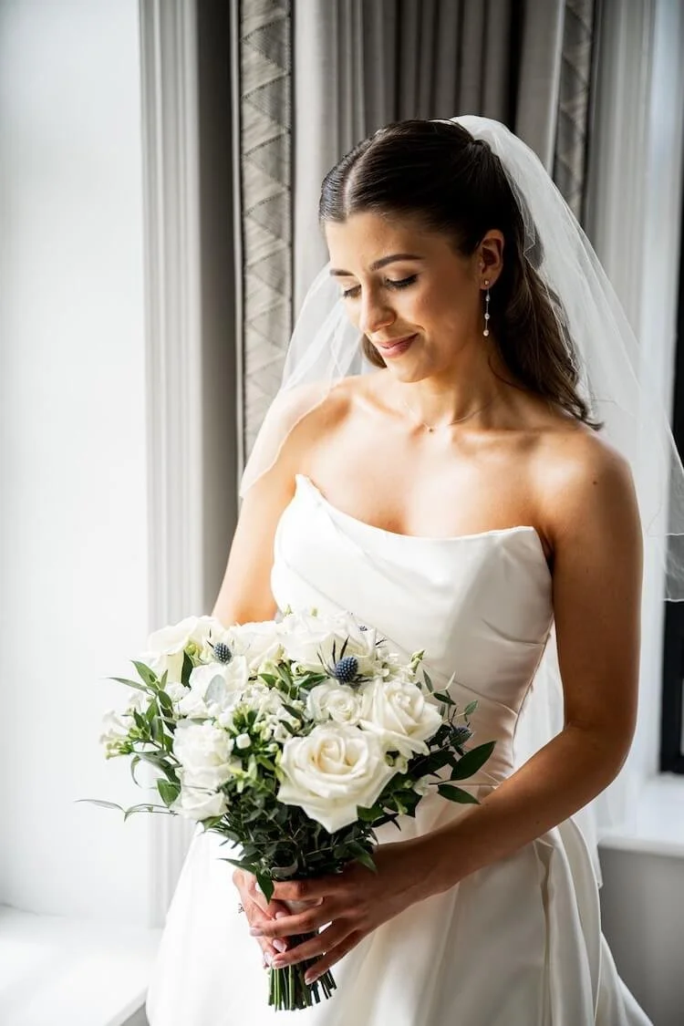 Bride in a white wedding dress holding a bouquet of white roses and greenery, with a veil and earrings, standing near window with curtains.