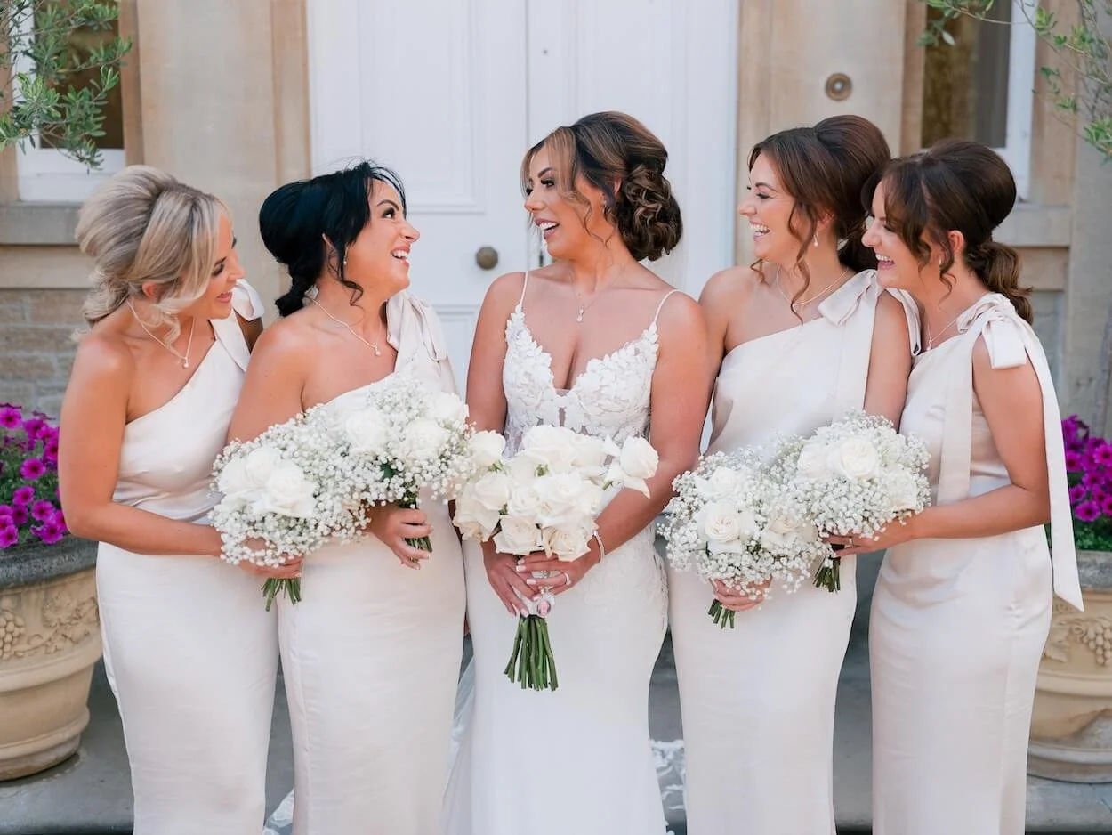A bride with four bridesmaids, all holding bouquets of white roses and gypsophila, standing outside a building with a white door background, all dressed in white and laughing.