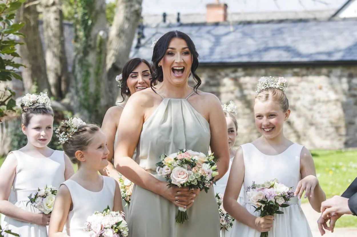 A bride in a white dress holding a bouquet of flowers while smiling happily. She is surrounded by young girls wearing white dresses and flower crowns, holding bouquets as well. The background features a stone building and trees, indicating an outdoor