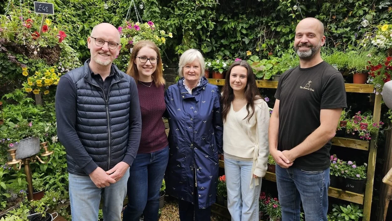 Group of five people standing outside against a background of garden plants and wooden shelving unit with garden plants for sale.