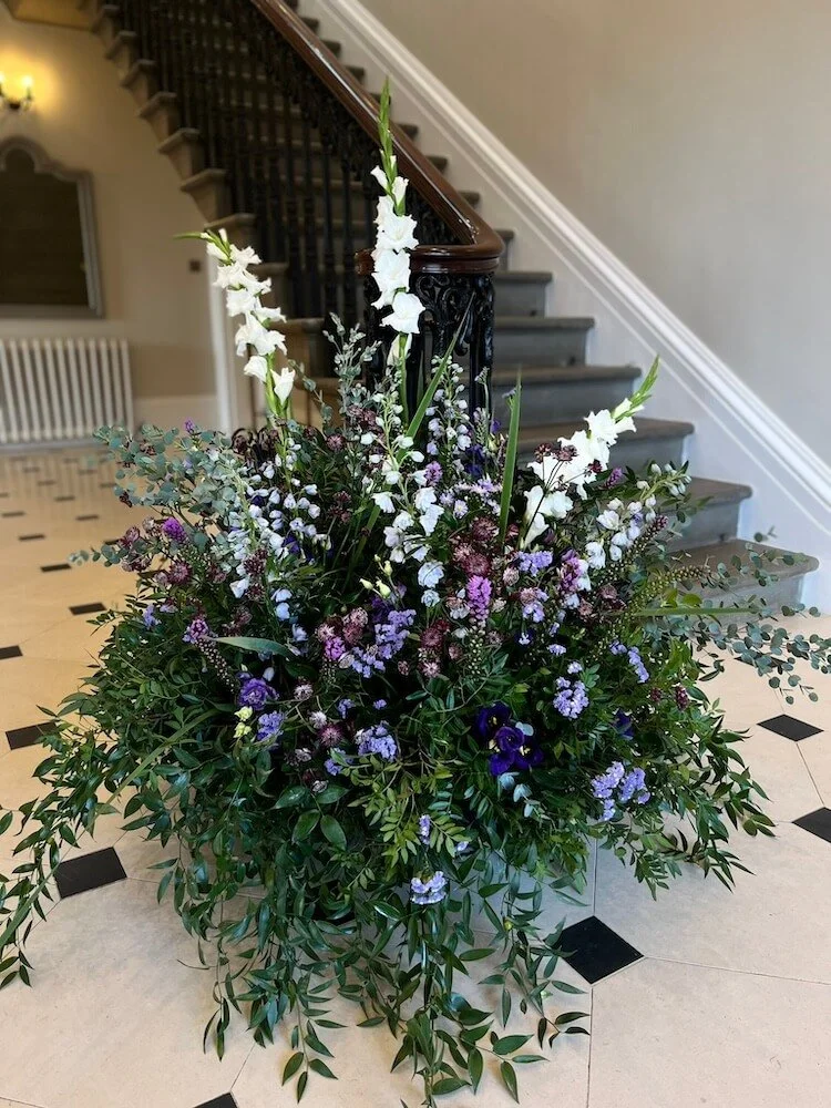 A large floral arrangement with white gladiolus, purple eustoma, and lavender-colored flowers, placed on a checkered floor near a staircase in an indoor setting.