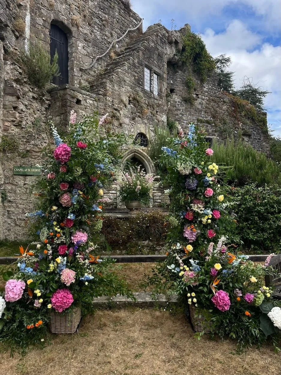 Colorful flower arrangements with pink, purple, yellow, and blue blooms on either side of a stone pathway leading to a small stone chapel with an arched entrance, set against a rustic stone wall with windows and greenery, under a partly cloudy sky.