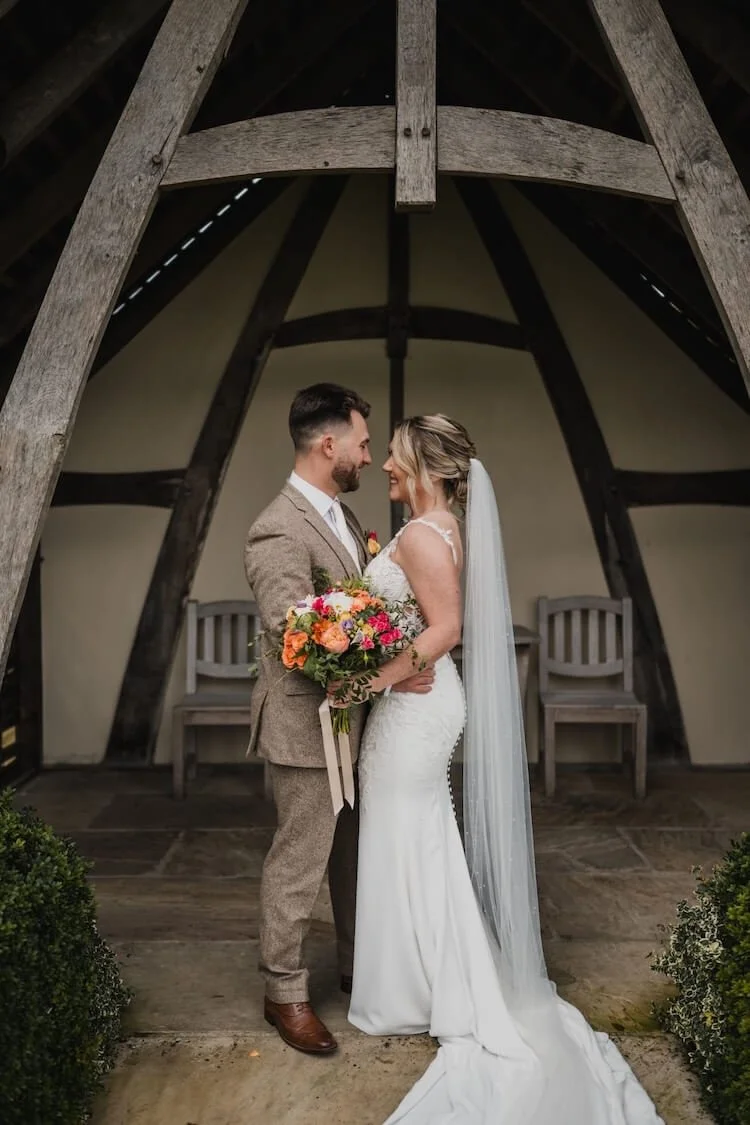 Bride and groom standing face to face under a rustic wooden structure, smiling at each other, holding a colourful bouquet bound with satin ribbon