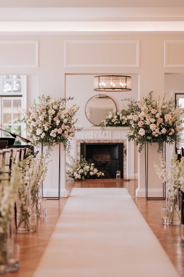 Elegant wedding ceremony setup with floral arrangements, a white aisle runner, and a fireplace with a circular mirror in a bright, airy room.