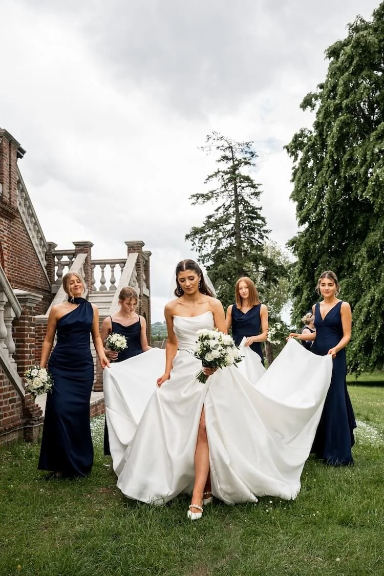 Bride in a white wedding dress walking on grass with five bridesmaids each holding up the bride's dress and wearing navy blue dresses, holding bouquets, near a brick building and large trees outdoors on a cloudy day.