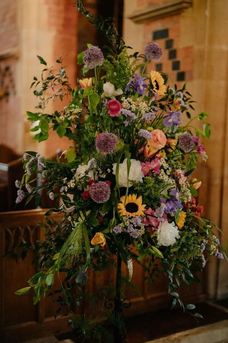 Colourful pedestal arrangement of various flowers, including sunflowers, roses, and purple blooms, arranged on a stand indoors.