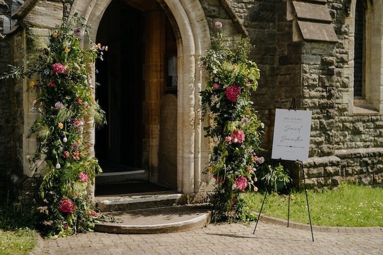 A loose, colourful broken floral arch framing the entrance of a stone church building, with a welcome sign on a stand nearby.