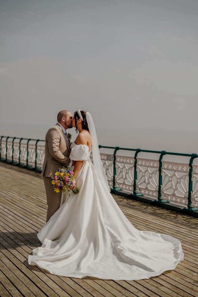 A bride and groom standing on a wooden deck by the water, sharing a kiss. The bride is in a white wedding dress holding a colourful bouquet, and the groom is in a beige suit.