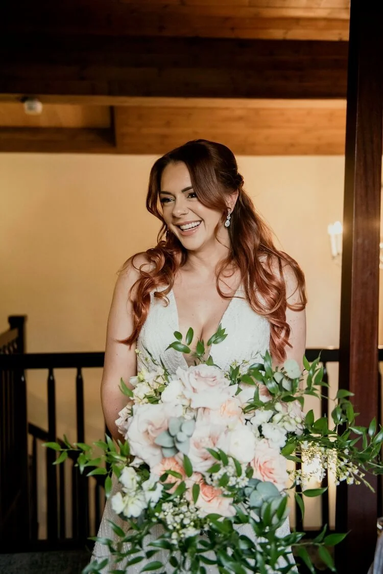 A bride with long, wavy red hair, wearing a white wedding dress, holding a large bouquet of light pink and white roses with green foliage, smiling and looking to the side indoors with wooden details and warm lighting.