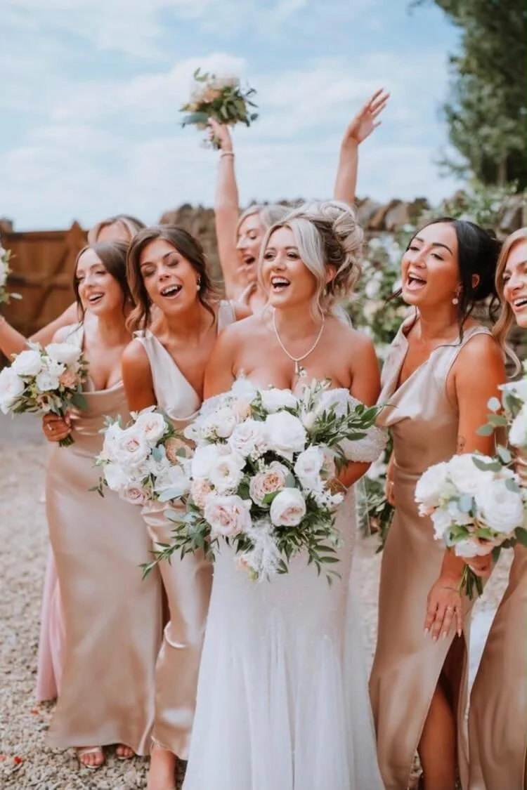 A bride and her bridesmaids celebrate outdoors, holding bouquets of flowers, smiling, with a wooden fence and trees in the background.