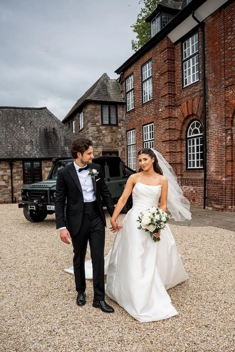 Bride and groom holding hands walking next to a brick building, with the bride wearing a white satin dress and holding a bouquet of white roses and greenery. Groom is dressed in a black tuxedo and there is a green off-road vehicle in the background
