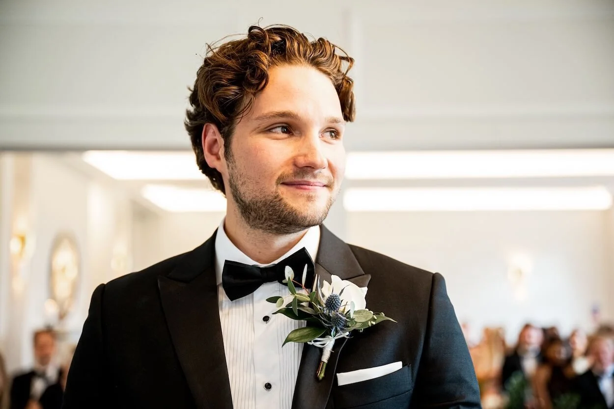 A man in a black tuxedo with a bow tie and a buttonhole, smiling and looking to the side.
