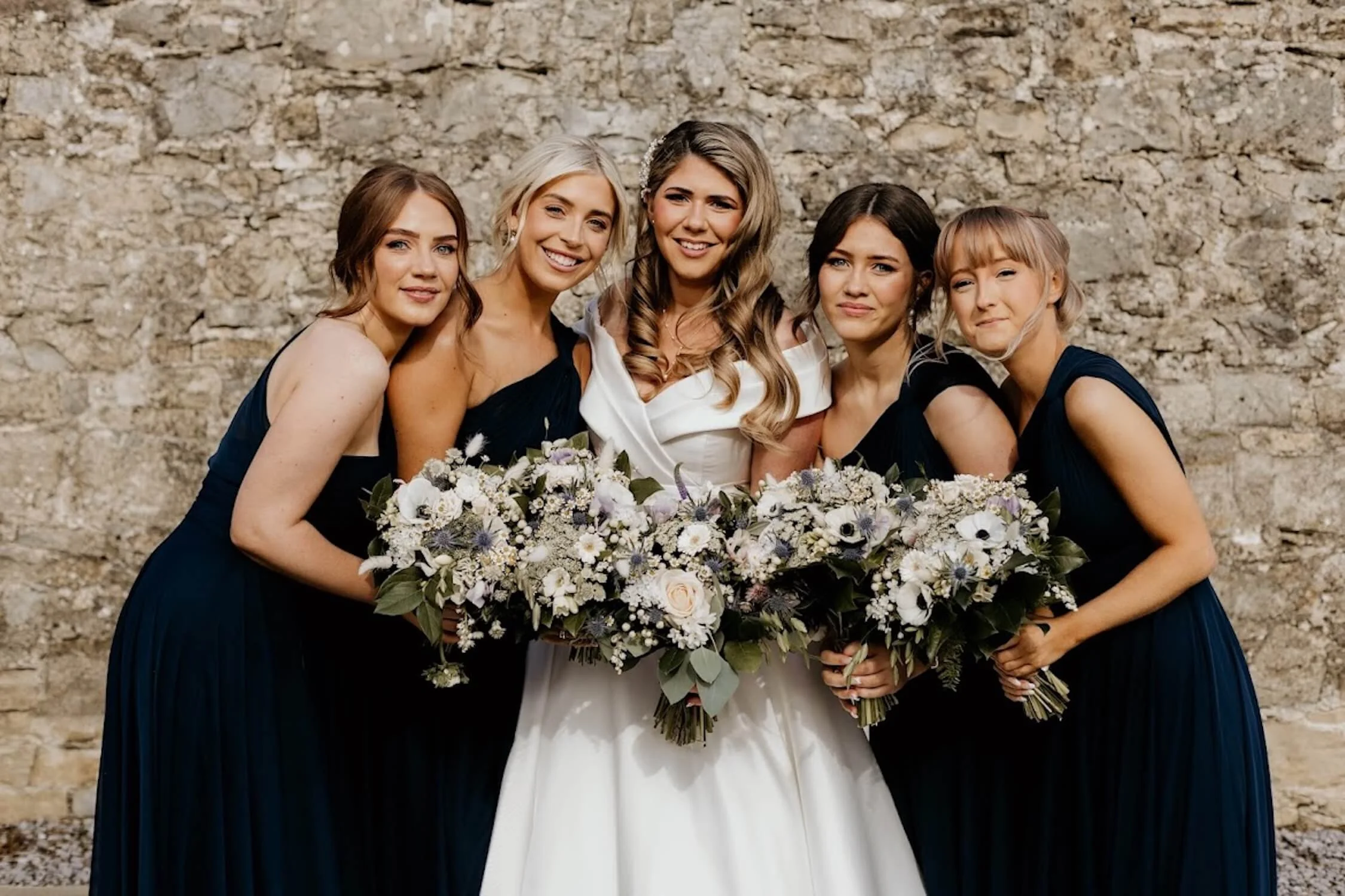 A group of five women dressed in formal gowns, standing close together outdoors in front of a stone wall, smiling, with one woman in the middle wearing a white wedding dress and holding a large bouquet of flowers.