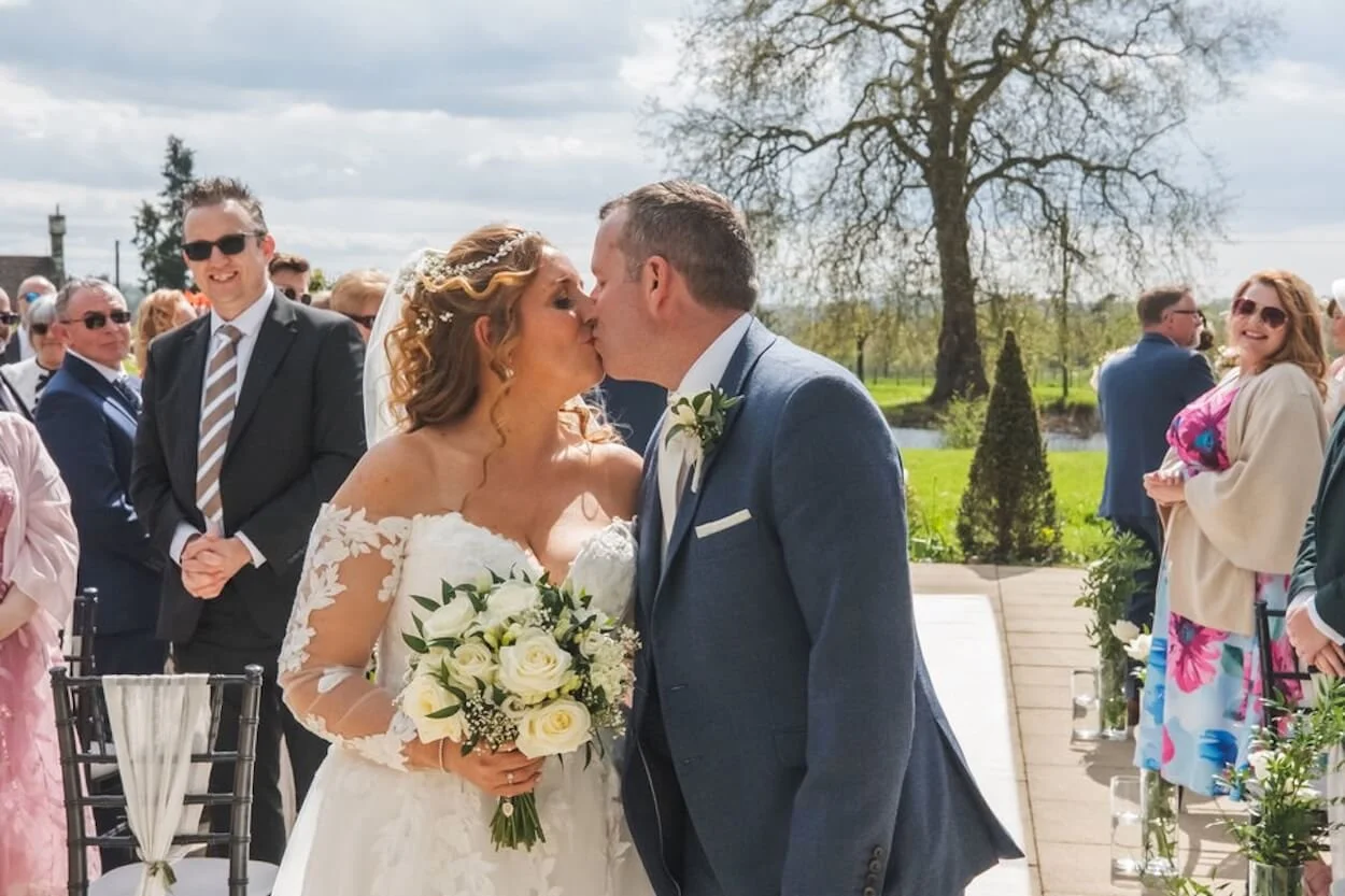 A bride and groom kiss during their outdoor wedding ceremony, surrounded by friends and family, with a large tree and greenery in the background.