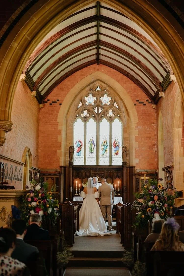 A bride and groom standing at the altar during their wedding ceremony inside a church with stained glass windows, wooden pews, and floral arrangements.