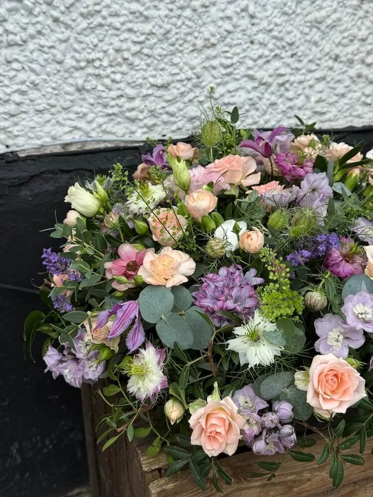 A colourful round posy in shades of lilac and peach, made up of various flowers including roses, lisianthus, clematis, and greenery placed on top of a wooden crate against a textured wall.