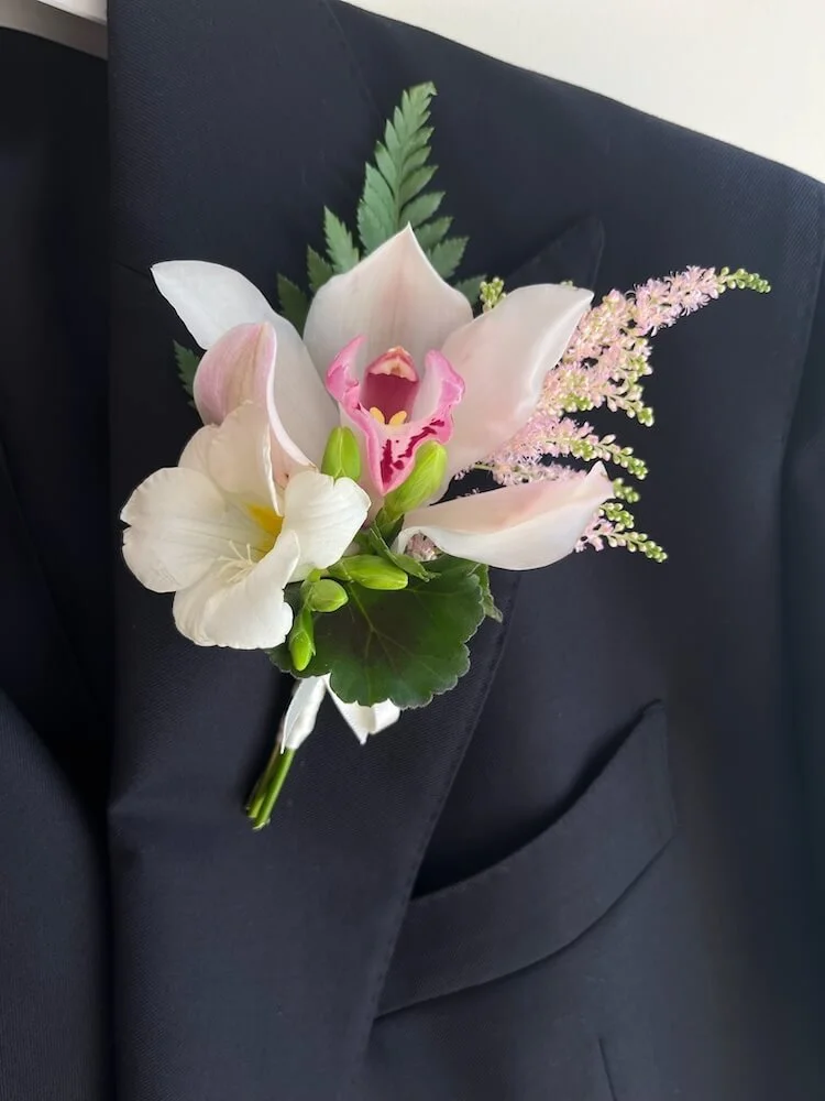A boutonniere with pink and white flowers, including a large white lily, a pink orchid, and small pink flowers with green leaves, pinned to a dark suit jacket.
