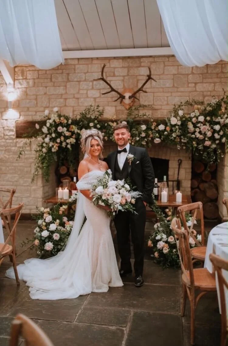 A bride and groom standing together in a wedding venue with floral decorations, a brick fireplace, and a mounted deer head with antlers on the wall.