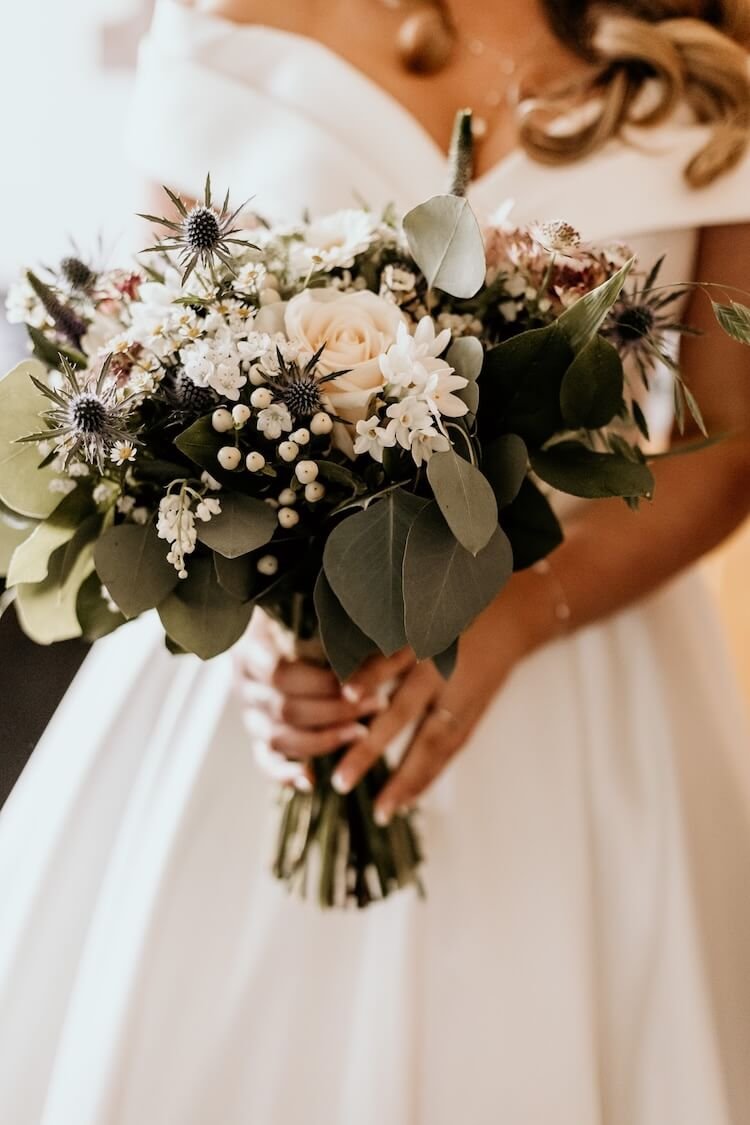 Close-up of a woman in a wedding dress holding a bouquet of white roses, thistles and green eucalyptus foliage..