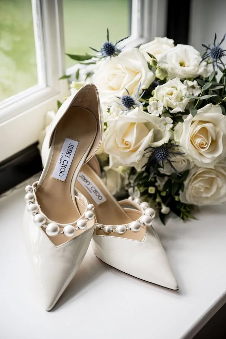 A pair of white pointed-toe high heels adorned with large pearl embellishments, placed on a windowsill next to a bouquet of white roses with accents of blue thistles.