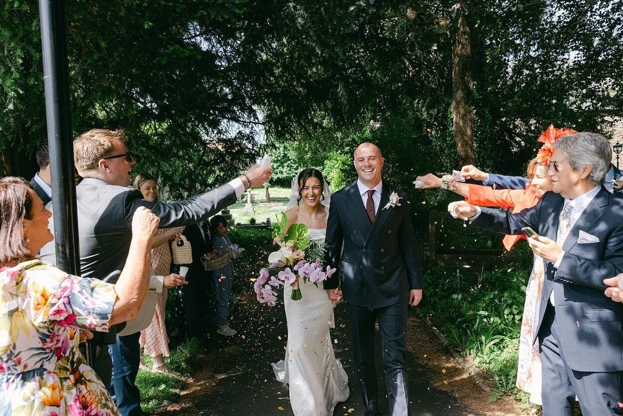 A bride and groom walking outdoors, holding hands, while guests throw flower petals at them. The bride is wearing a white wedding gown with a bouquet of flowers, and the groom is in a dark suit with a boutonniere. The setting is a lush, green garden 