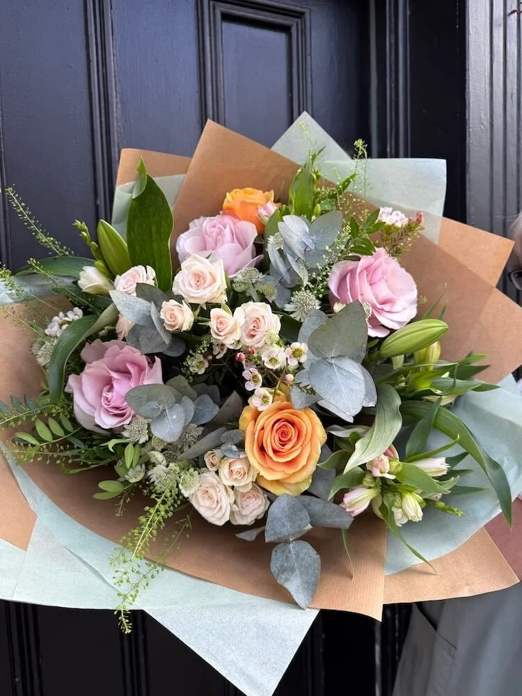 A bouquet of mixed flowers including pink roses, peach roses, lilies, and greenery wrapped in brown and sage green tissue paper.