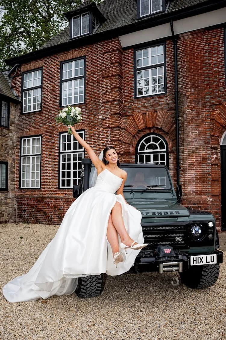 Bride in wedding dress sitting on top of the front of a green Land Rover Defender, holding a bouquet of white roses with a happy expression, in front of a brick house.