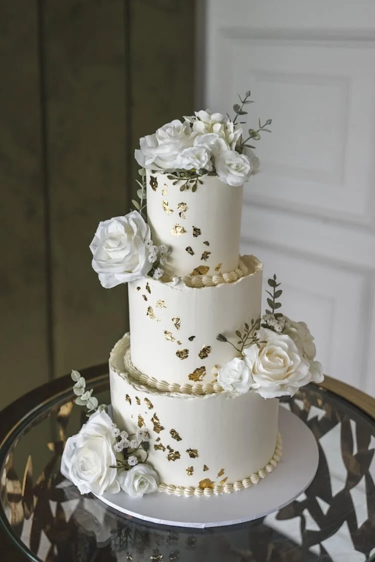 Three-tier white wedding cake decorated with white roses, green leaves, small white flowers, and gold leaf accents, displayed on a glass table.
