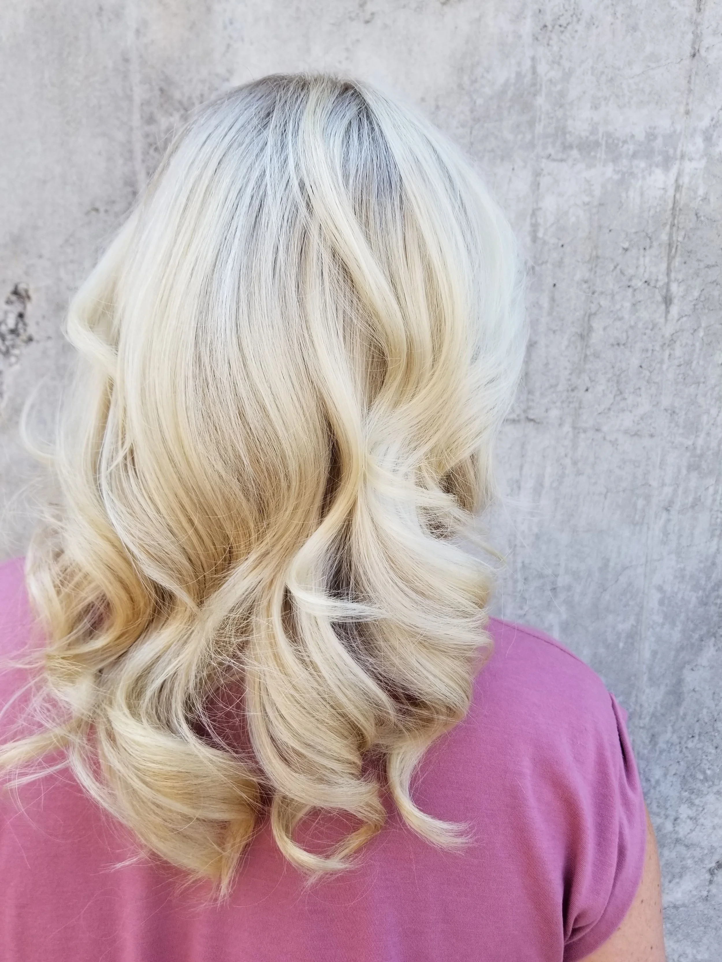 Close-up image of blonde, wavy hair of a woman wearing a purple top, against a gray concrete wall background.