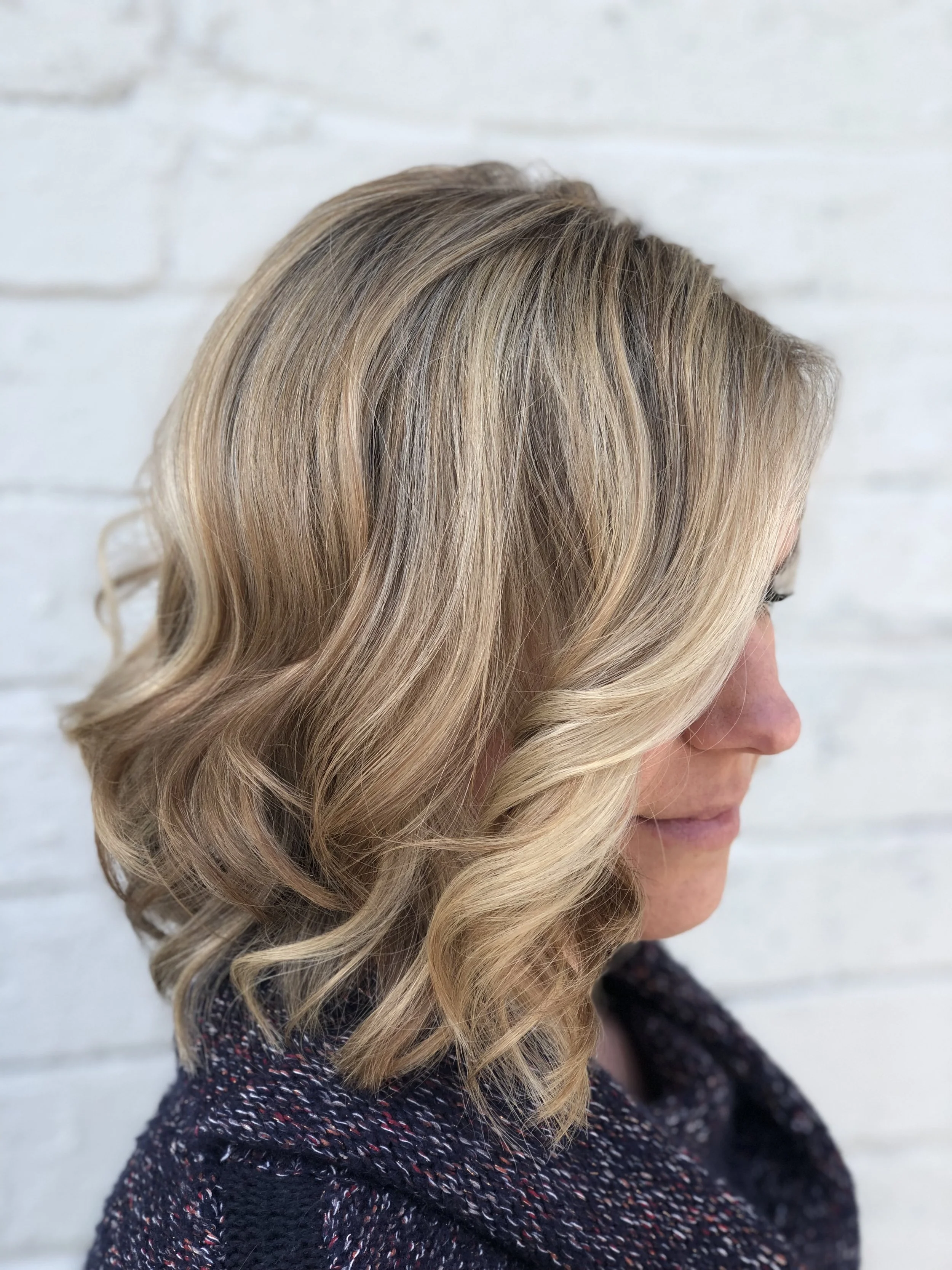 Side view of a woman with blonde, wavy hair in front of a white brick wall.