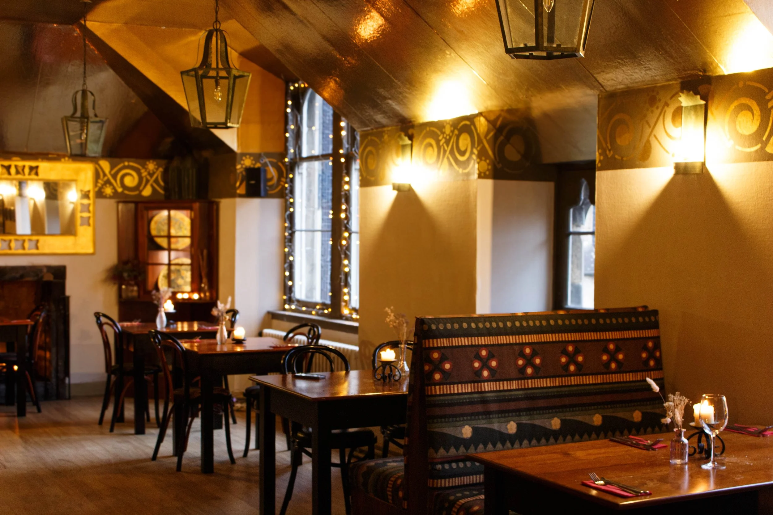 Cozy restaurant interior with wooden tables set with glasses, candles, and cutlery, decorated with hanging lanterns, string lights, and a painted wall with patterns.