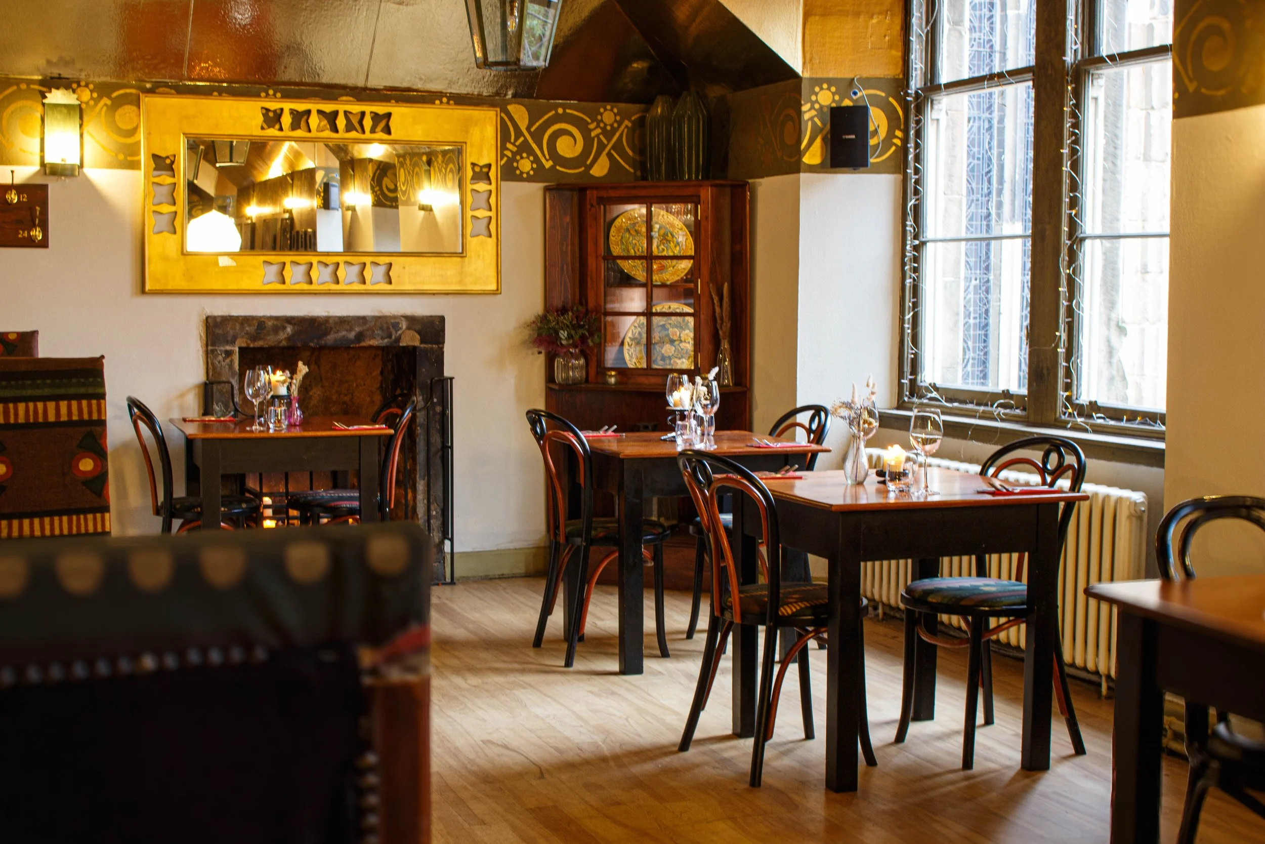 Interior of a cozy restaurant with wooden tables set with glasses, candles, and utensils. Decor includes a large gold-framed mirror, a wooden display cabinet with decorative plates, and windows with string lights. Warm lighting enhances a welcoming atmosphere.