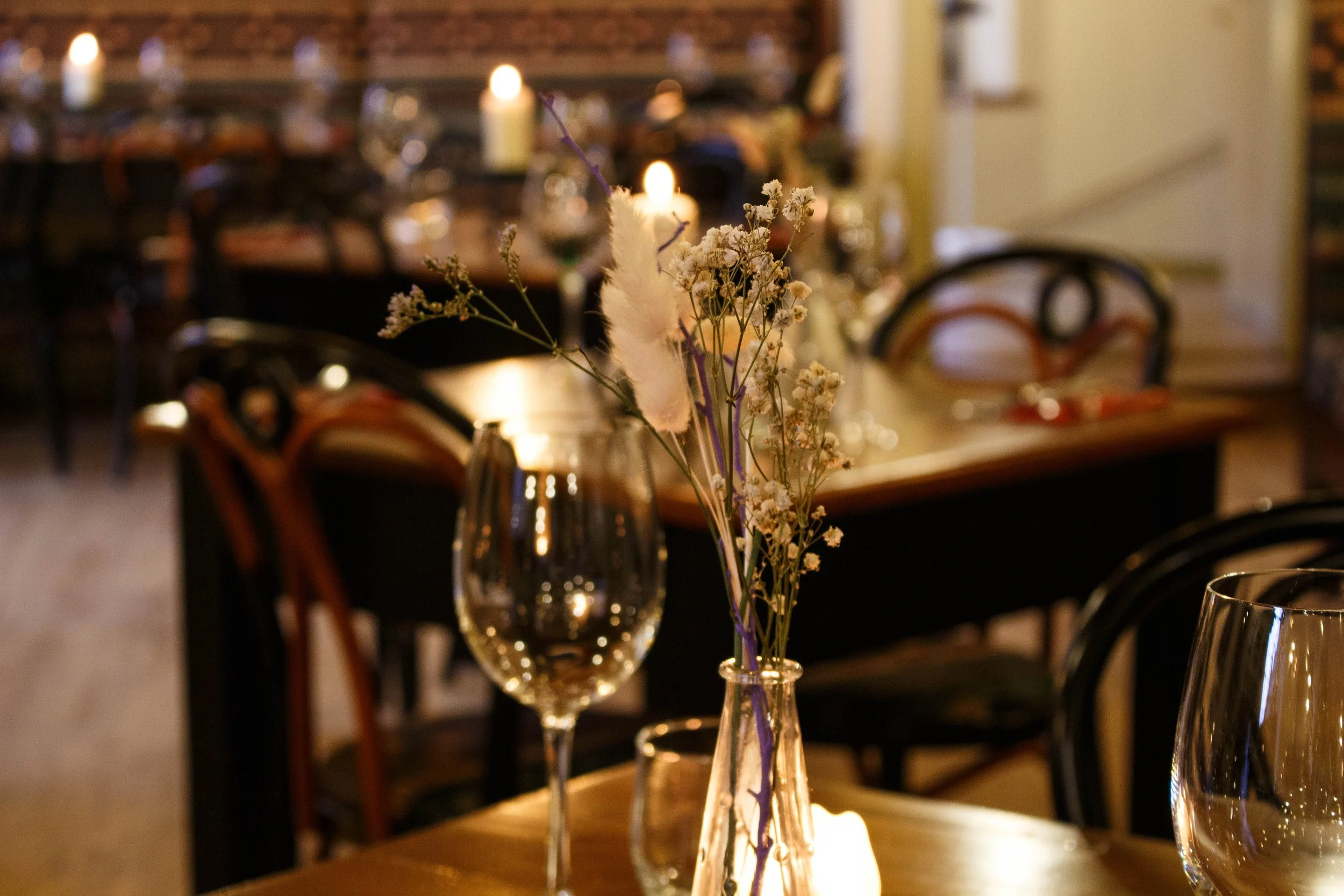 A table centerpiece with dried flowers in a glass vase, lit by candlelight, in a dimly lit dining area with blurred chairs and tables in the background.