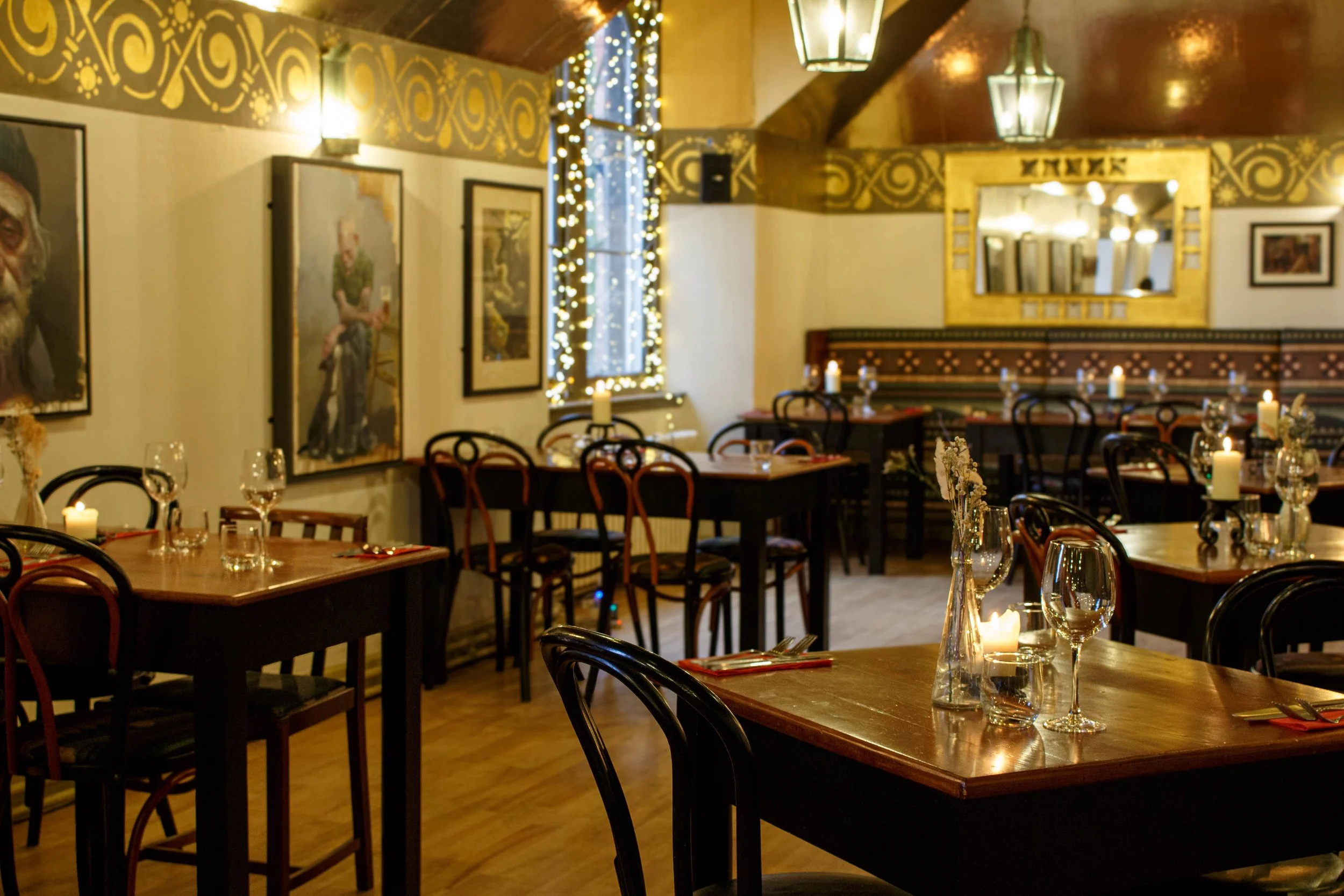 A cozy restaurant interior decorated with string lights, candles, paintings, and ornate gold and black details, set for dining with tables, wine glasses, and tableware.