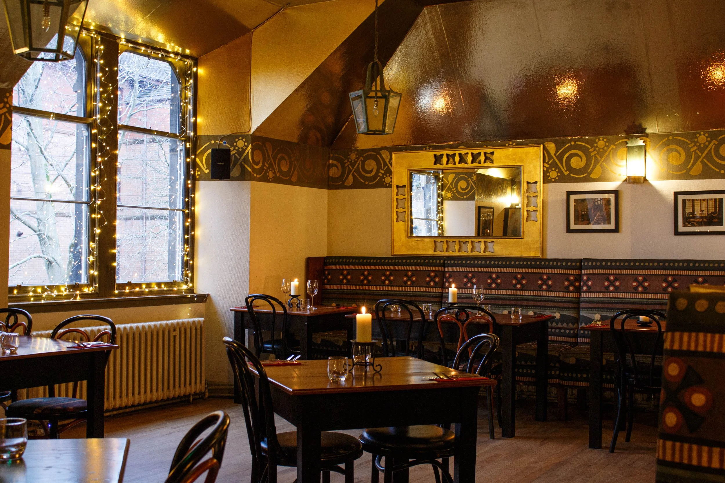 Interior of a cozy restaurant with warm lighting, decorated with string lights around a large window showing trees outside, framed pictures on the walls, a golden mirror, and candles on tables.